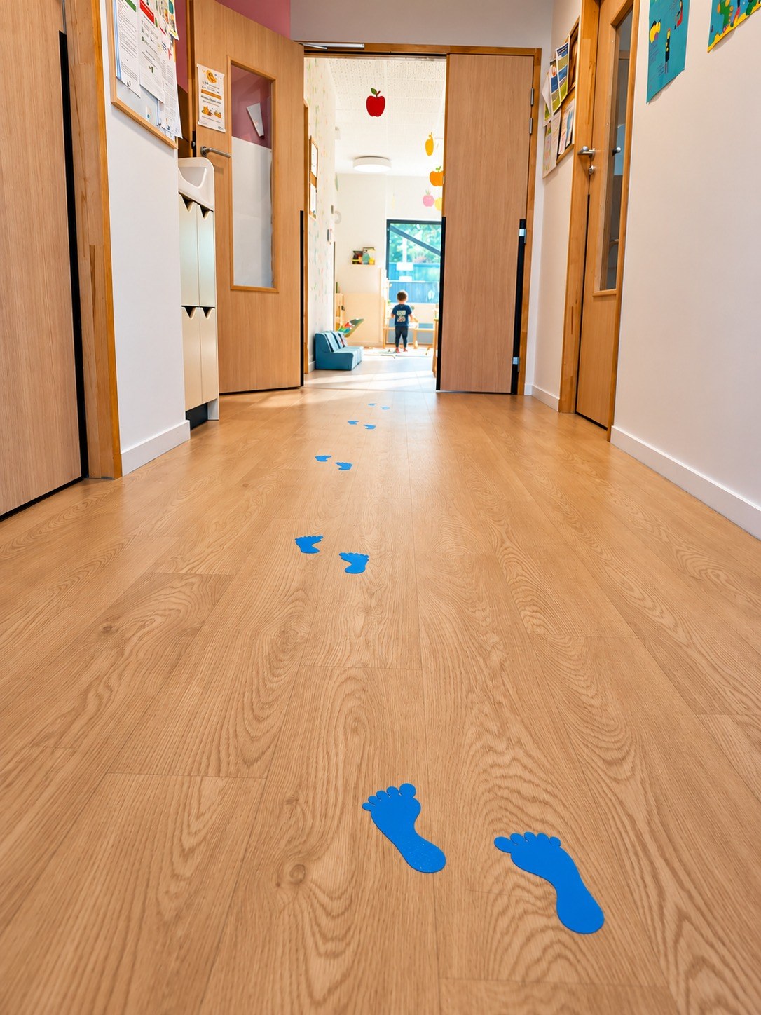 Couloir de la crèche avec empreintes de pas bleues menant à la salle de vie, enfant vu de dos au fond